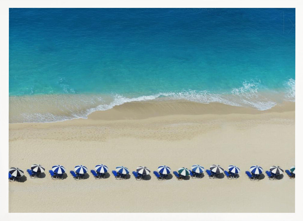 An aerial view of a pristine sandy beach with vibrant turquoise ocean waves gently lapping the shore. A long, neat row of blue and white striped beach umbrellas stands along the sand, creating a serene and orderly coastal scene, all enclosed within a silver frame. Artwork