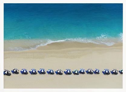An aerial view of a pristine sandy beach with vibrant turquoise ocean waves gently lapping the shore. A long, neat row of blue and white striped beach umbrellas stands along the sand, creating a serene and orderly coastal scene, all enclosed within a silver frame. Artwork