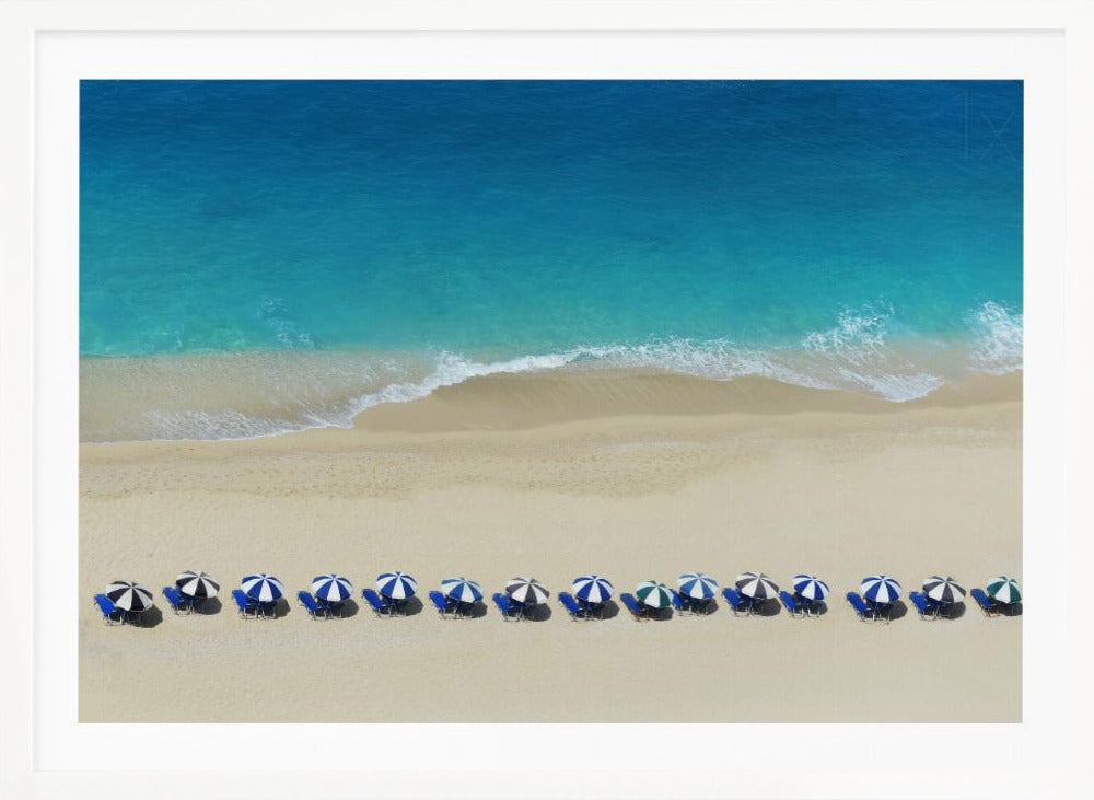 An aerial view of a pristine sandy beach with vibrant turquoise ocean waves gently lapping the shore. A long, neat row of blue and white striped beach umbrellas stands along the sand, creating a serene and orderly coastal scene, all enclosed within a silver frame. Artwork