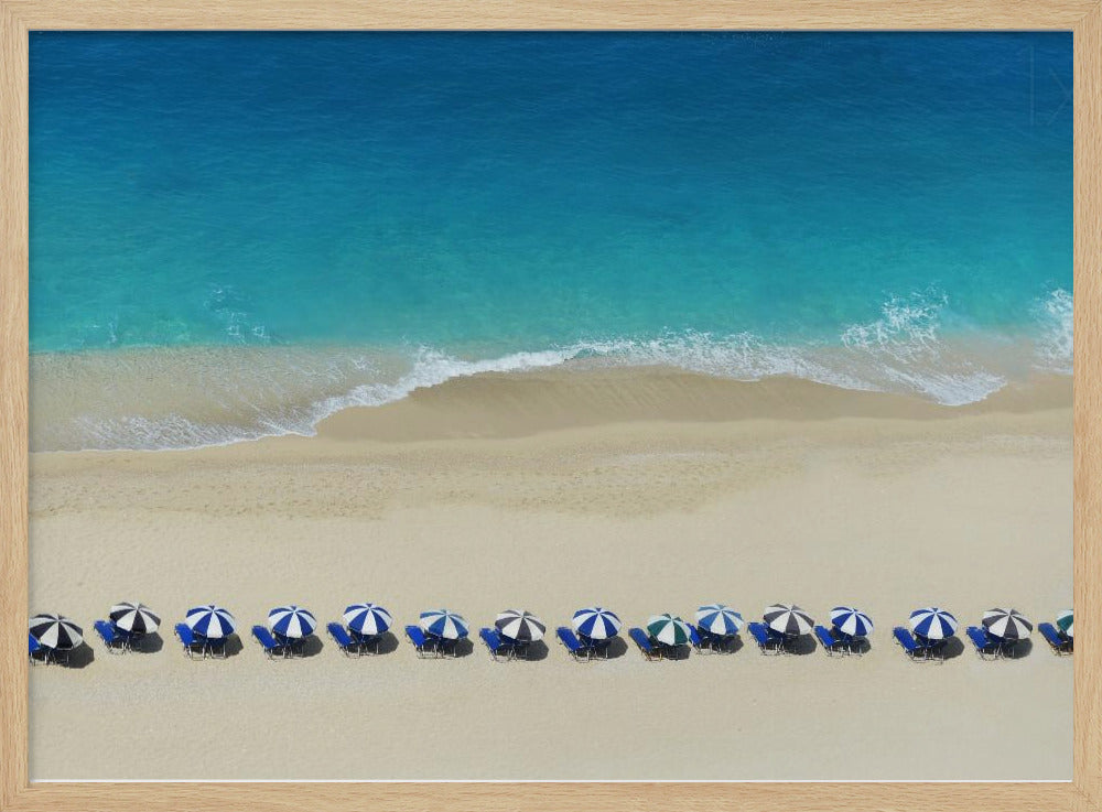 An aerial view of a pristine sandy beach with vibrant turquoise ocean waves gently lapping the shore. A long, neat row of blue and white striped beach umbrellas stands along the sand, creating a serene and orderly coastal scene, all enclosed within a silver frame. Artwork