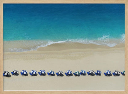 An aerial view of a pristine sandy beach with vibrant turquoise ocean waves gently lapping the shore. A long, neat row of blue and white striped beach umbrellas stands along the sand, creating a serene and orderly coastal scene, all enclosed within a silver frame. Artwork