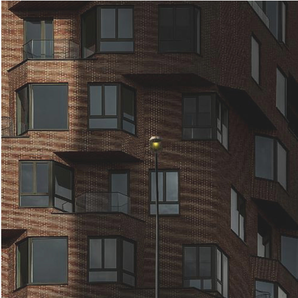 A close-up photograph of a modern red-brick apartment building, framed in light wood. A sharp shadow line divides the facade, with a lit streetlamp providing a warm yellow glow in the shaded area. Decor