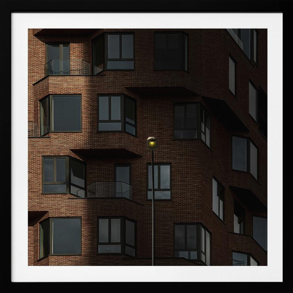 A close-up photograph of a modern red-brick apartment building, framed in light wood. A sharp shadow line divides the facade, with a lit streetlamp providing a warm yellow glow in the shaded area. Decor