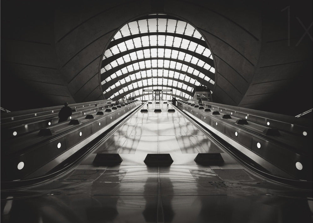 A dramatic low-angle, black and white photograph of the escalators at Canary Wharf station. The perspective looks up the sleek, reflective escalators towards a massive, arched, grid-patterned window at the far end, creating strong leading lines and a sense of scale within the modern, curved architecture. Decor