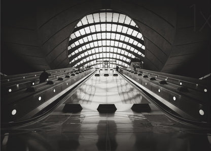 A dramatic low-angle, black and white photograph of the escalators at Canary Wharf station. The perspective looks up the sleek, reflective escalators towards a massive, arched, grid-patterned window at the far end, creating strong leading lines and a sense of scale within the modern, curved architecture. Decor