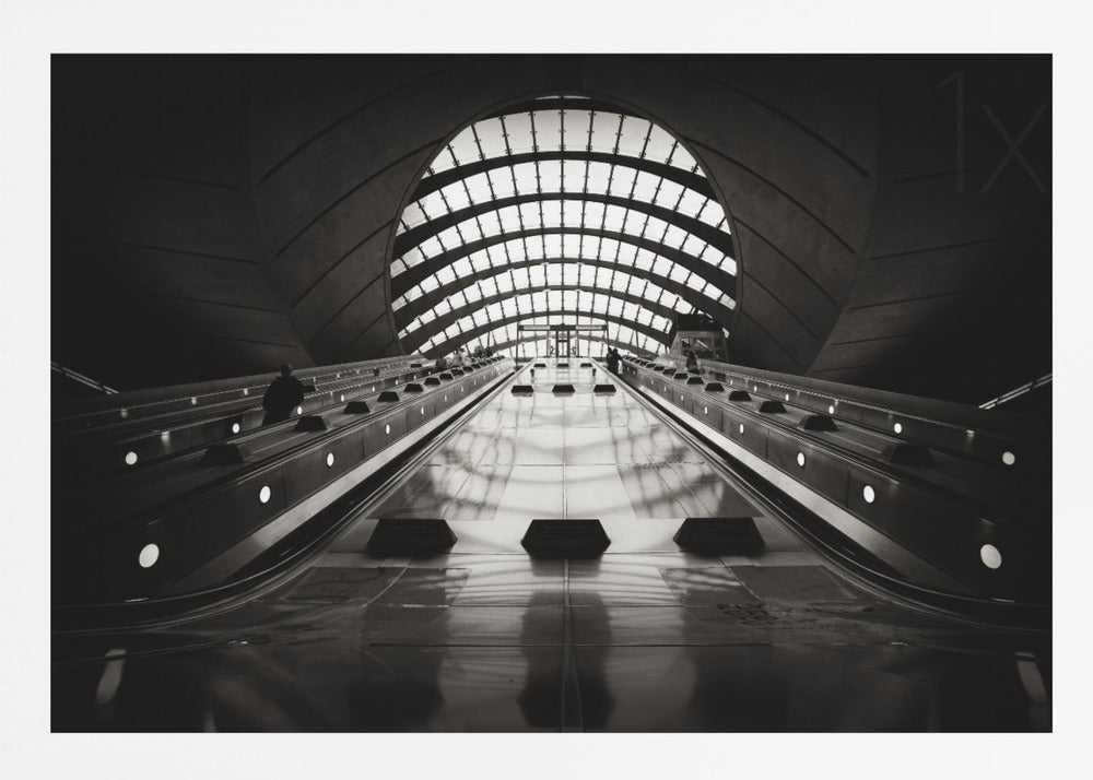 A dramatic low-angle, black and white photograph of the escalators at Canary Wharf station. The perspective looks up the sleek, reflective escalators towards a massive, arched, grid-patterned window at the far end, creating strong leading lines and a sense of scale within the modern, curved architecture. Decor