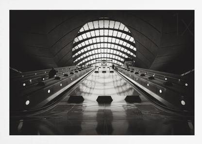 A dramatic low-angle, black and white photograph of the escalators at Canary Wharf station. The perspective looks up the sleek, reflective escalators towards a massive, arched, grid-patterned window at the far end, creating strong leading lines and a sense of scale within the modern, curved architecture. Decor