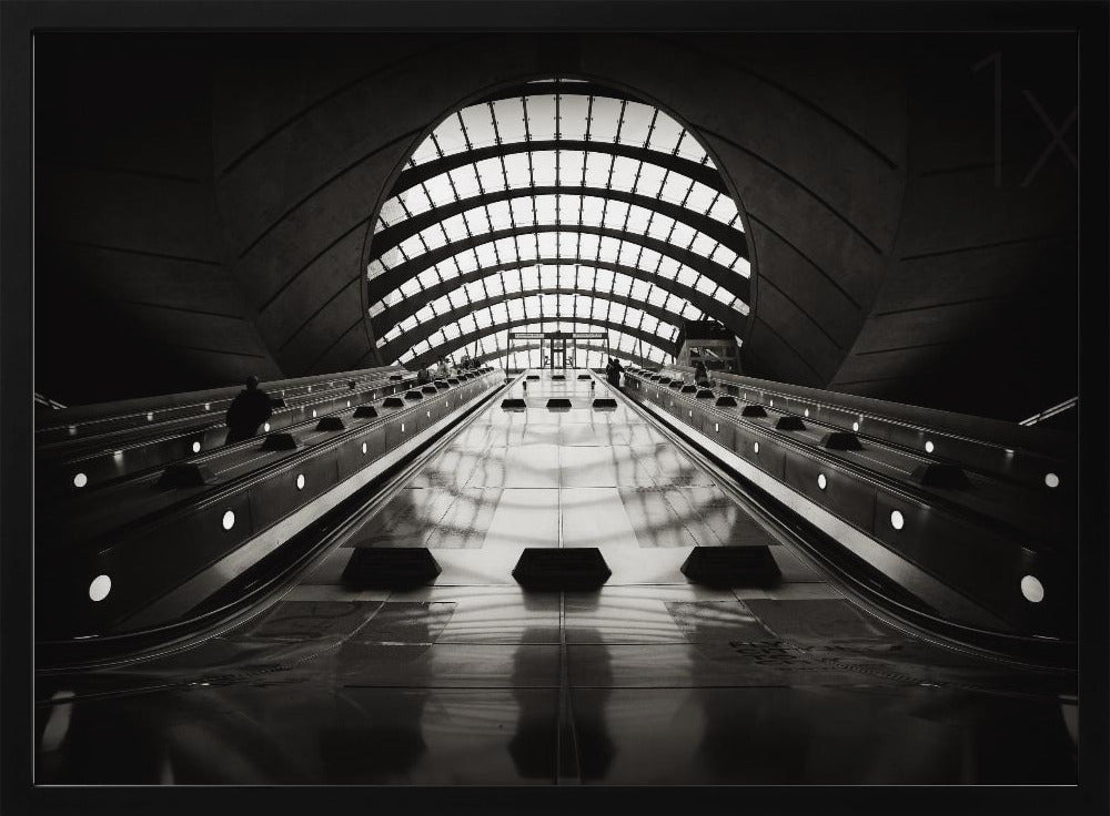A dramatic low-angle, black and white photograph of the escalators at Canary Wharf station. The perspective looks up the sleek, reflective escalators towards a massive, arched, grid-patterned window at the far end, creating strong leading lines and a sense of scale within the modern, curved architecture. Decor