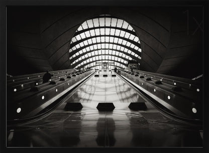 A dramatic low-angle, black and white photograph of the escalators at Canary Wharf station. The perspective looks up the sleek, reflective escalators towards a massive, arched, grid-patterned window at the far end, creating strong leading lines and a sense of scale within the modern, curved architecture. Decor