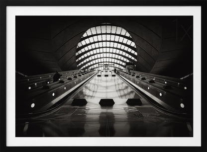 A dramatic low-angle, black and white photograph of the escalators at Canary Wharf station. The perspective looks up the sleek, reflective escalators towards a massive, arched, grid-patterned window at the far end, creating strong leading lines and a sense of scale within the modern, curved architecture. Decor