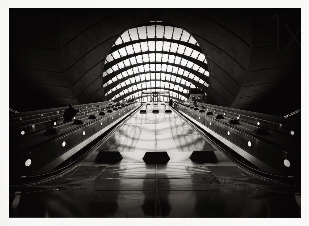 A dramatic low-angle, black and white photograph of the escalators at Canary Wharf station. The perspective looks up the sleek, reflective escalators towards a massive, arched, grid-patterned window at the far end, creating strong leading lines and a sense of scale within the modern, curved architecture. Decor
