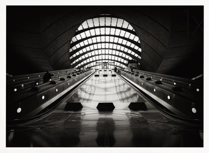 A dramatic low-angle, black and white photograph of the escalators at Canary Wharf station. The perspective looks up the sleek, reflective escalators towards a massive, arched, grid-patterned window at the far end, creating strong leading lines and a sense of scale within the modern, curved architecture. Decor