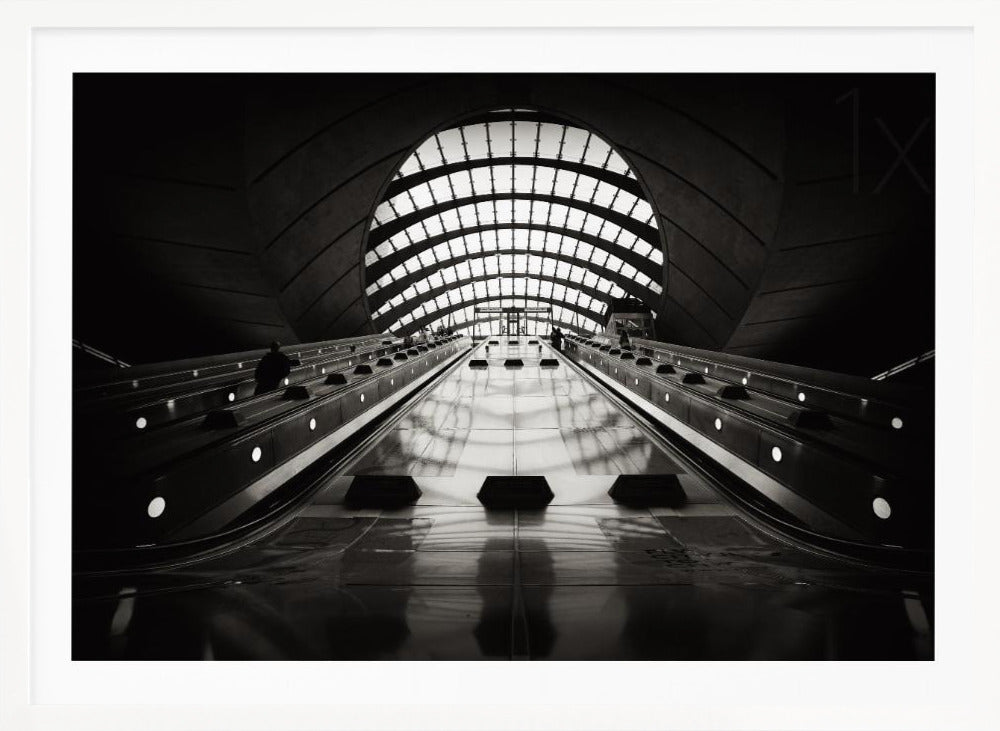 A dramatic low-angle, black and white photograph of the escalators at Canary Wharf station. The perspective looks up the sleek, reflective escalators towards a massive, arched, grid-patterned window at the far end, creating strong leading lines and a sense of scale within the modern, curved architecture. Decor
