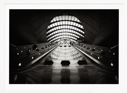 A dramatic low-angle, black and white photograph of the escalators at Canary Wharf station. The perspective looks up the sleek, reflective escalators towards a massive, arched, grid-patterned window at the far end, creating strong leading lines and a sense of scale within the modern, curved architecture. Decor