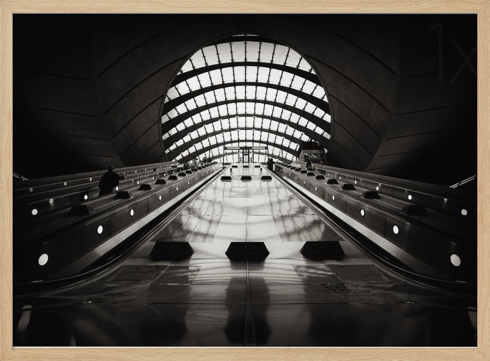 A dramatic low-angle, black and white photograph of the escalators at Canary Wharf station. The perspective looks up the sleek, reflective escalators towards a massive, arched, grid-patterned window at the far end, creating strong leading lines and a sense of scale within the modern, curved architecture. Decor