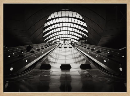 A dramatic low-angle, black and white photograph of the escalators at Canary Wharf station. The perspective looks up the sleek, reflective escalators towards a massive, arched, grid-patterned window at the far end, creating strong leading lines and a sense of scale within the modern, curved architecture. Decor