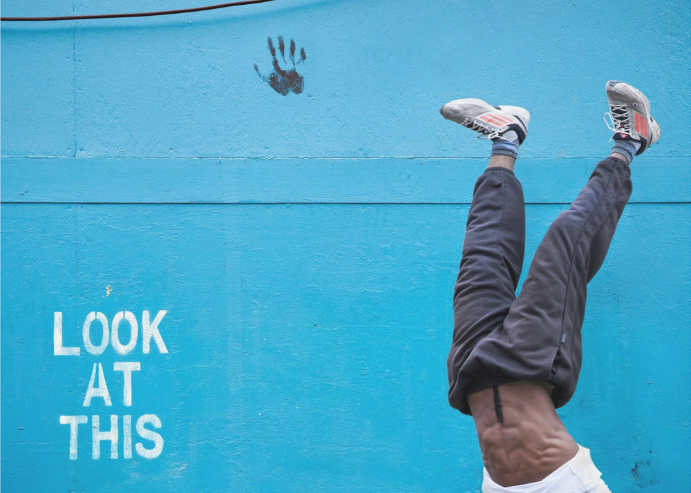 A person's lower body doing a handstand against a bright blue wall with the stenciled words 'LOOK AT THIS' in white. The person wears dark pants and sneakers, and their muscular abdomen is visible. A black handprint is on the wall above the text. The artwork is presented in a silver frame. Print