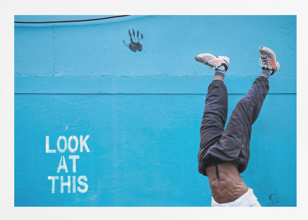 A person's lower body doing a handstand against a bright blue wall with the stenciled words 'LOOK AT THIS' in white. The person wears dark pants and sneakers, and their muscular abdomen is visible. A black handprint is on the wall above the text. The artwork is presented in a silver frame. Print