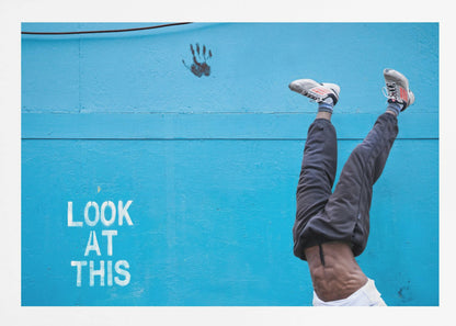 A person's lower body doing a handstand against a bright blue wall with the stenciled words 'LOOK AT THIS' in white. The person wears dark pants and sneakers, and their muscular abdomen is visible. A black handprint is on the wall above the text. The artwork is presented in a silver frame. Print
