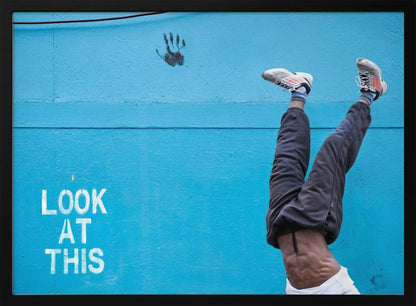 A person's lower body doing a handstand against a bright blue wall with the stenciled words 'LOOK AT THIS' in white. The person wears dark pants and sneakers, and their muscular abdomen is visible. A black handprint is on the wall above the text. The artwork is presented in a silver frame. Print