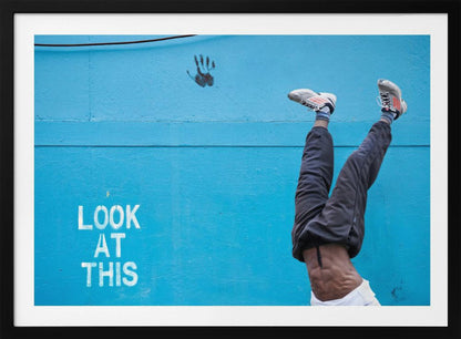 A person's lower body doing a handstand against a bright blue wall with the stenciled words 'LOOK AT THIS' in white. The person wears dark pants and sneakers, and their muscular abdomen is visible. A black handprint is on the wall above the text. The artwork is presented in a silver frame. Print