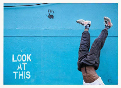 A person's lower body doing a handstand against a bright blue wall with the stenciled words 'LOOK AT THIS' in white. The person wears dark pants and sneakers, and their muscular abdomen is visible. A black handprint is on the wall above the text. The artwork is presented in a silver frame. Print
