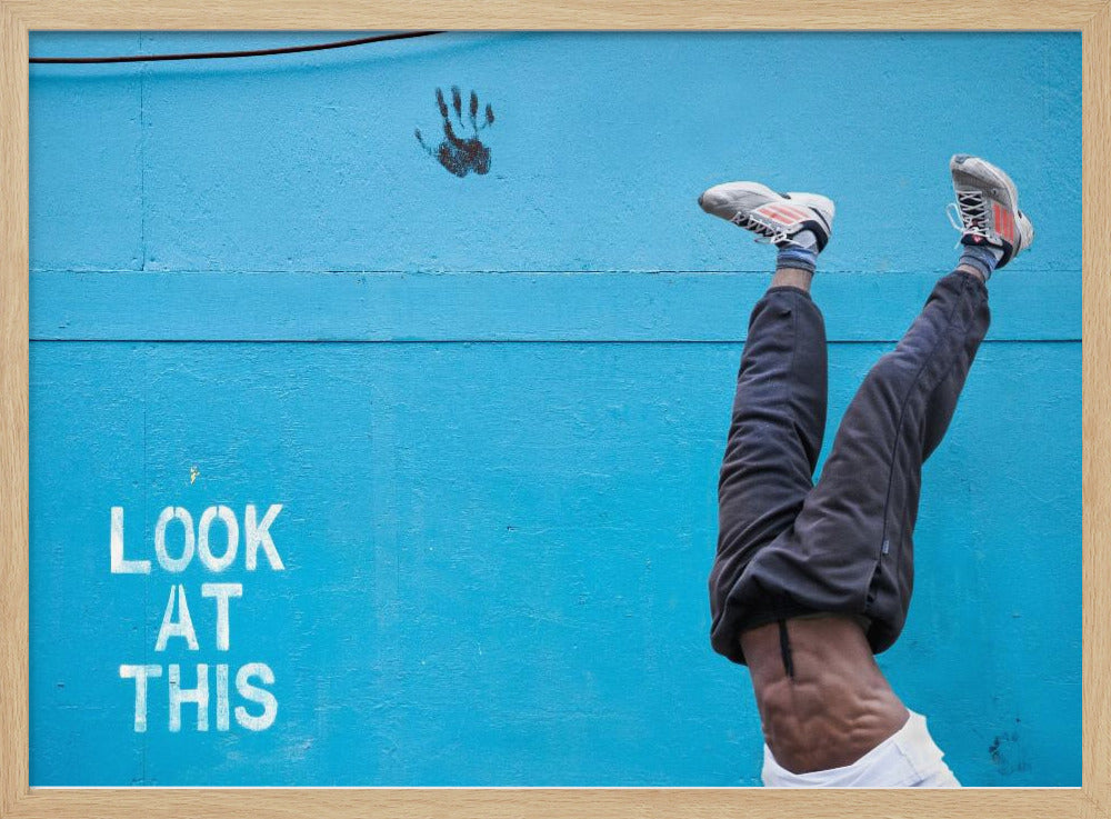 A person's lower body doing a handstand against a bright blue wall with the stenciled words 'LOOK AT THIS' in white. The person wears dark pants and sneakers, and their muscular abdomen is visible. A black handprint is on the wall above the text. The artwork is presented in a silver frame. Print