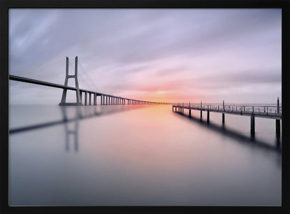 A serene long-exposure photograph of a modern cable-stayed bridge and a pier stretching over mirror-like water at sunset. The sky is filled with soft, streaky clouds in hues of pink and purple, reflected on the calm water's surface, all enclosed in a silver frame. Decor