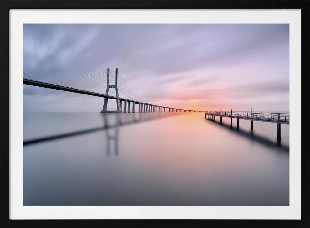 A serene long-exposure photograph of a modern cable-stayed bridge and a pier stretching over mirror-like water at sunset. The sky is filled with soft, streaky clouds in hues of pink and purple, reflected on the calm water's surface, all enclosed in a silver frame. Decor