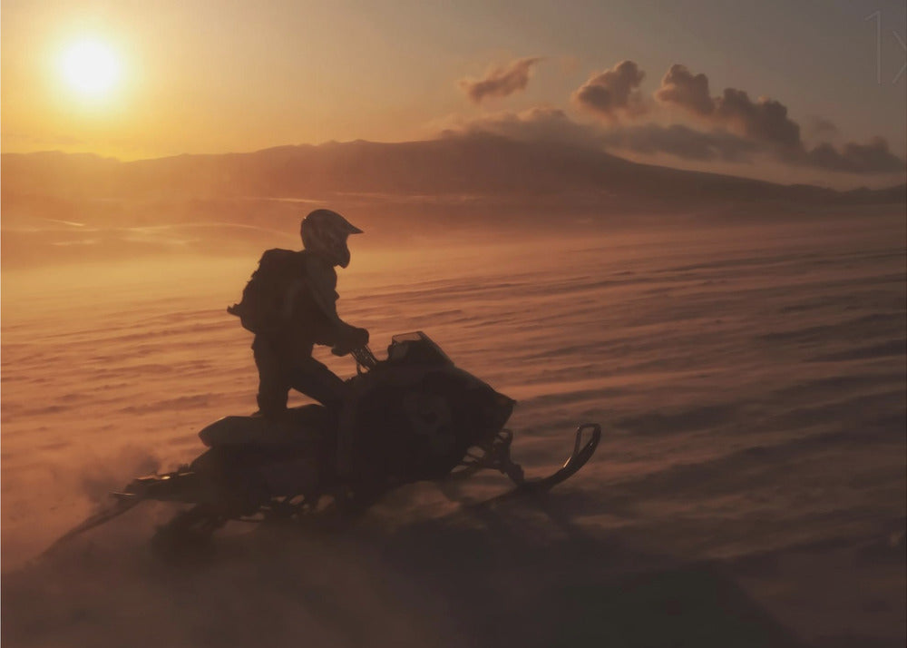 A silhouette of a person wearing a helmet and backpack stands on a snowmobile in a vast, snowy landscape during a golden sunset. The wind blows snow across the ground, creating a hazy, atmospheric effect, with mountains visible in the distance under a warm sky. Wall Art