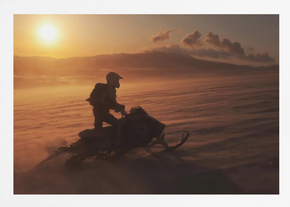 A silhouette of a person wearing a helmet and backpack stands on a snowmobile in a vast, snowy landscape during a golden sunset. The wind blows snow across the ground, creating a hazy, atmospheric effect, with mountains visible in the distance under a warm sky. Wall Art