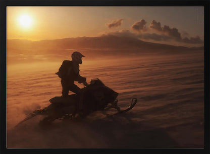 A silhouette of a person wearing a helmet and backpack stands on a snowmobile in a vast, snowy landscape during a golden sunset. The wind blows snow across the ground, creating a hazy, atmospheric effect, with mountains visible in the distance under a warm sky. Wall Art