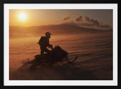 A silhouette of a person wearing a helmet and backpack stands on a snowmobile in a vast, snowy landscape during a golden sunset. The wind blows snow across the ground, creating a hazy, atmospheric effect, with mountains visible in the distance under a warm sky. Wall Art