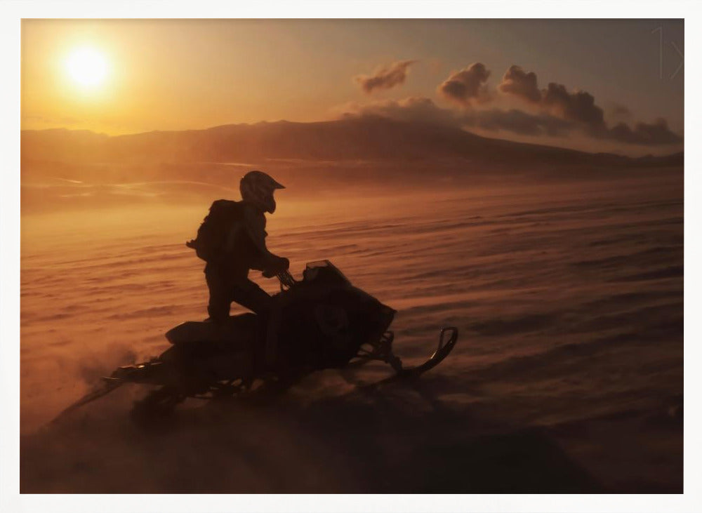 A silhouette of a person wearing a helmet and backpack stands on a snowmobile in a vast, snowy landscape during a golden sunset. The wind blows snow across the ground, creating a hazy, atmospheric effect, with mountains visible in the distance under a warm sky. Wall Art
