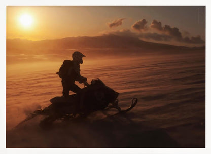 A silhouette of a person wearing a helmet and backpack stands on a snowmobile in a vast, snowy landscape during a golden sunset. The wind blows snow across the ground, creating a hazy, atmospheric effect, with mountains visible in the distance under a warm sky. Wall Art