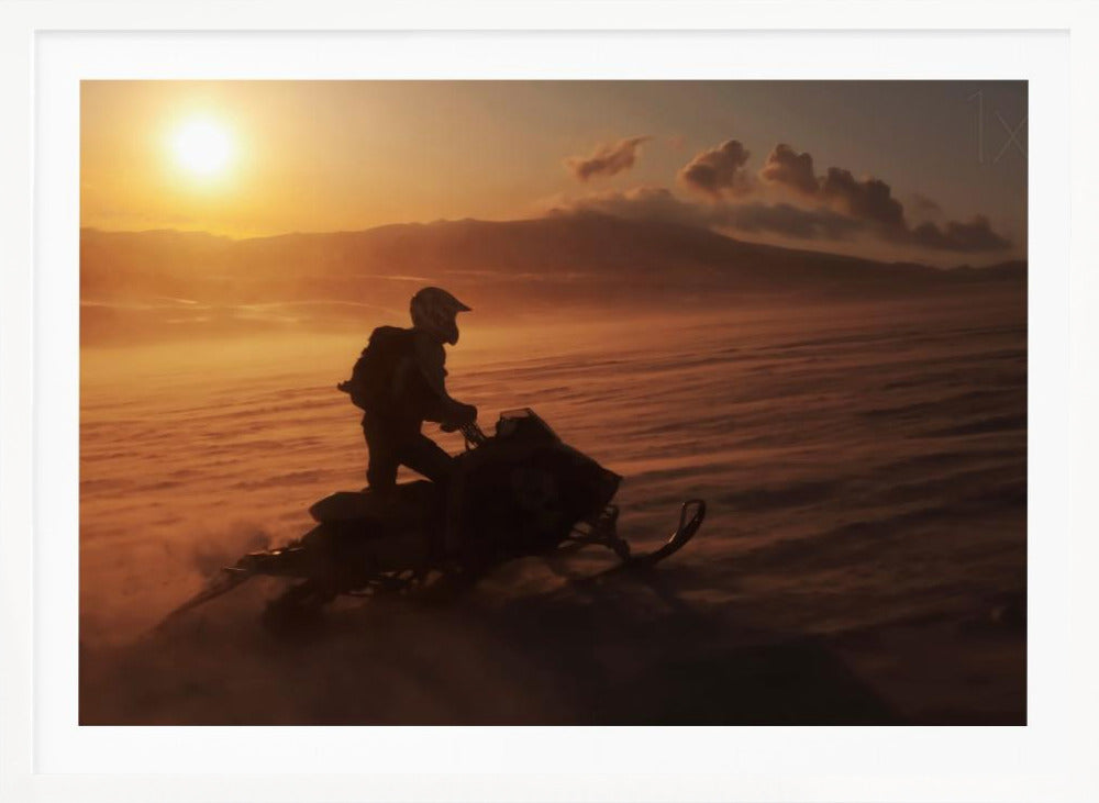 A silhouette of a person wearing a helmet and backpack stands on a snowmobile in a vast, snowy landscape during a golden sunset. The wind blows snow across the ground, creating a hazy, atmospheric effect, with mountains visible in the distance under a warm sky. Wall Art