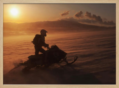 A silhouette of a person wearing a helmet and backpack stands on a snowmobile in a vast, snowy landscape during a golden sunset. The wind blows snow across the ground, creating a hazy, atmospheric effect, with mountains visible in the distance under a warm sky. Wall Art