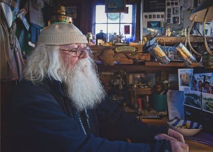 A profile portrait of an elderly man with a long, full white beard and hair, wearing glasses and a cream-colored knit beanie. He sits indoors in a warmly lit, cluttered room that looks like a cabin or a pub, with his hands clasped in front of him, gazing thoughtfully to the side. Poster