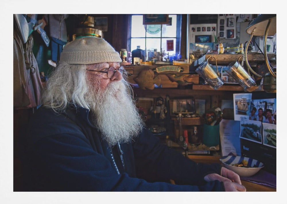 A profile portrait of an elderly man with a long, full white beard and hair, wearing glasses and a cream-colored knit beanie. He sits indoors in a warmly lit, cluttered room that looks like a cabin or a pub, with his hands clasped in front of him, gazing thoughtfully to the side. Poster