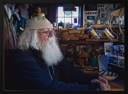 A profile portrait of an elderly man with a long, full white beard and hair, wearing glasses and a cream-colored knit beanie. He sits indoors in a warmly lit, cluttered room that looks like a cabin or a pub, with his hands clasped in front of him, gazing thoughtfully to the side. Poster