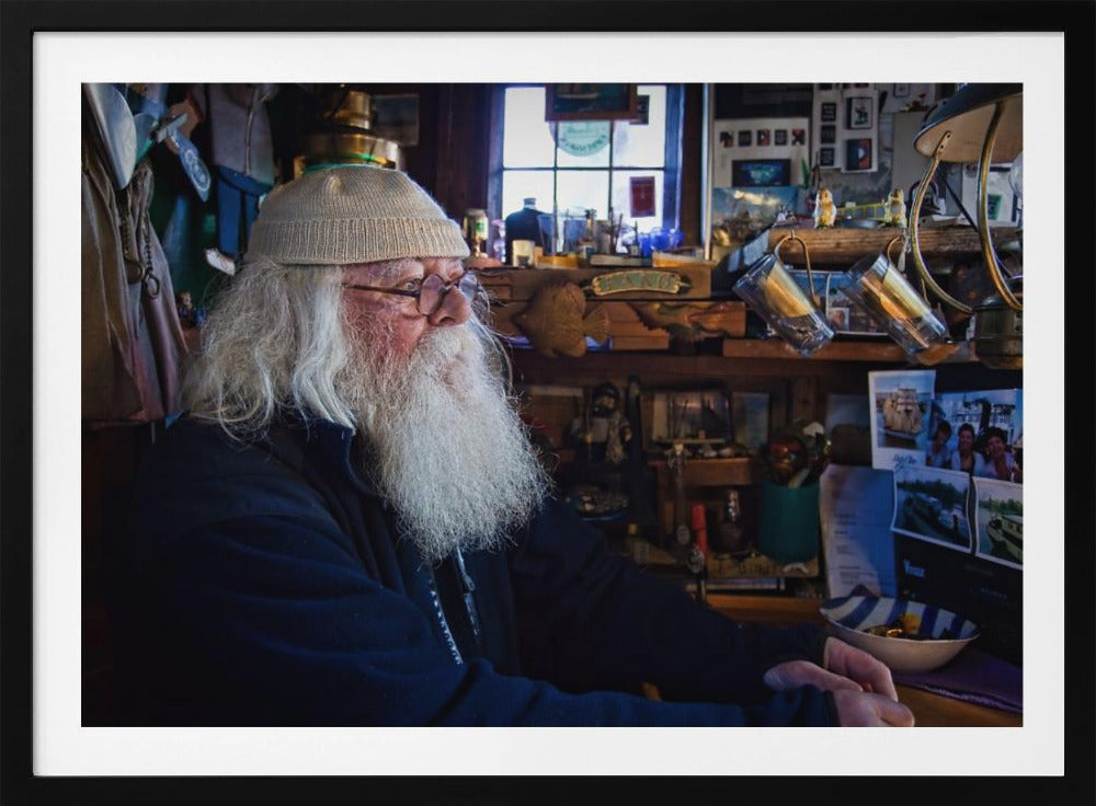 A profile portrait of an elderly man with a long, full white beard and hair, wearing glasses and a cream-colored knit beanie. He sits indoors in a warmly lit, cluttered room that looks like a cabin or a pub, with his hands clasped in front of him, gazing thoughtfully to the side. Poster