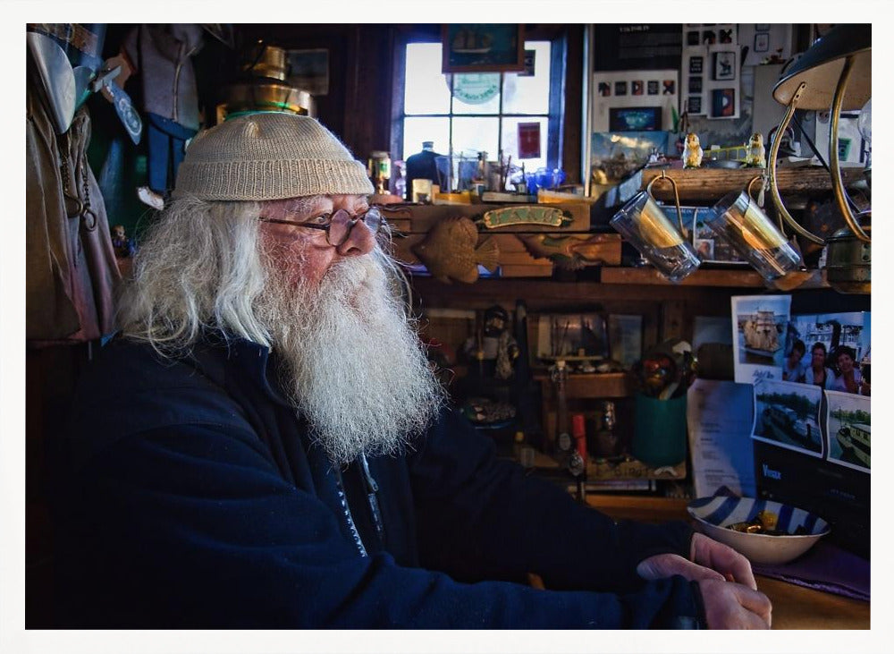 A profile portrait of an elderly man with a long, full white beard and hair, wearing glasses and a cream-colored knit beanie. He sits indoors in a warmly lit, cluttered room that looks like a cabin or a pub, with his hands clasped in front of him, gazing thoughtfully to the side. Poster