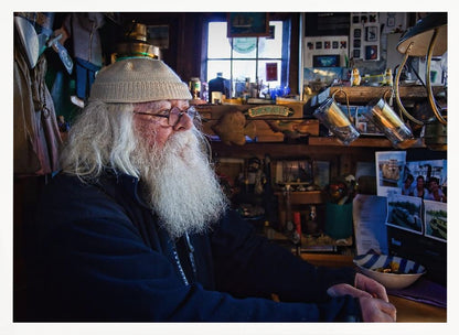 A profile portrait of an elderly man with a long, full white beard and hair, wearing glasses and a cream-colored knit beanie. He sits indoors in a warmly lit, cluttered room that looks like a cabin or a pub, with his hands clasped in front of him, gazing thoughtfully to the side. Poster