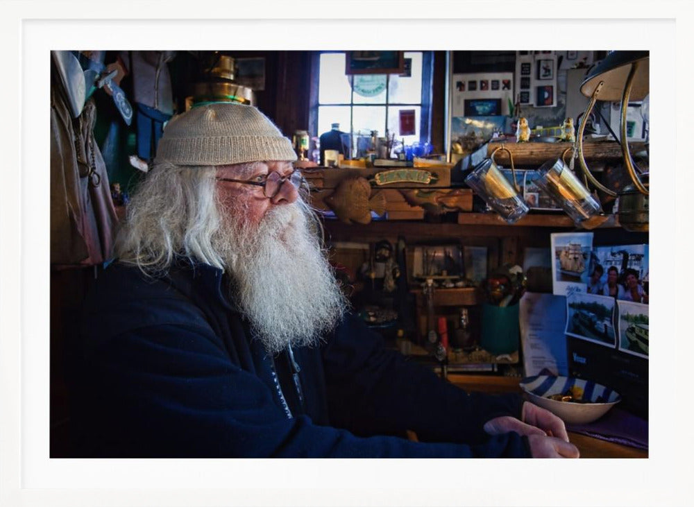 A profile portrait of an elderly man with a long, full white beard and hair, wearing glasses and a cream-colored knit beanie. He sits indoors in a warmly lit, cluttered room that looks like a cabin or a pub, with his hands clasped in front of him, gazing thoughtfully to the side. Poster