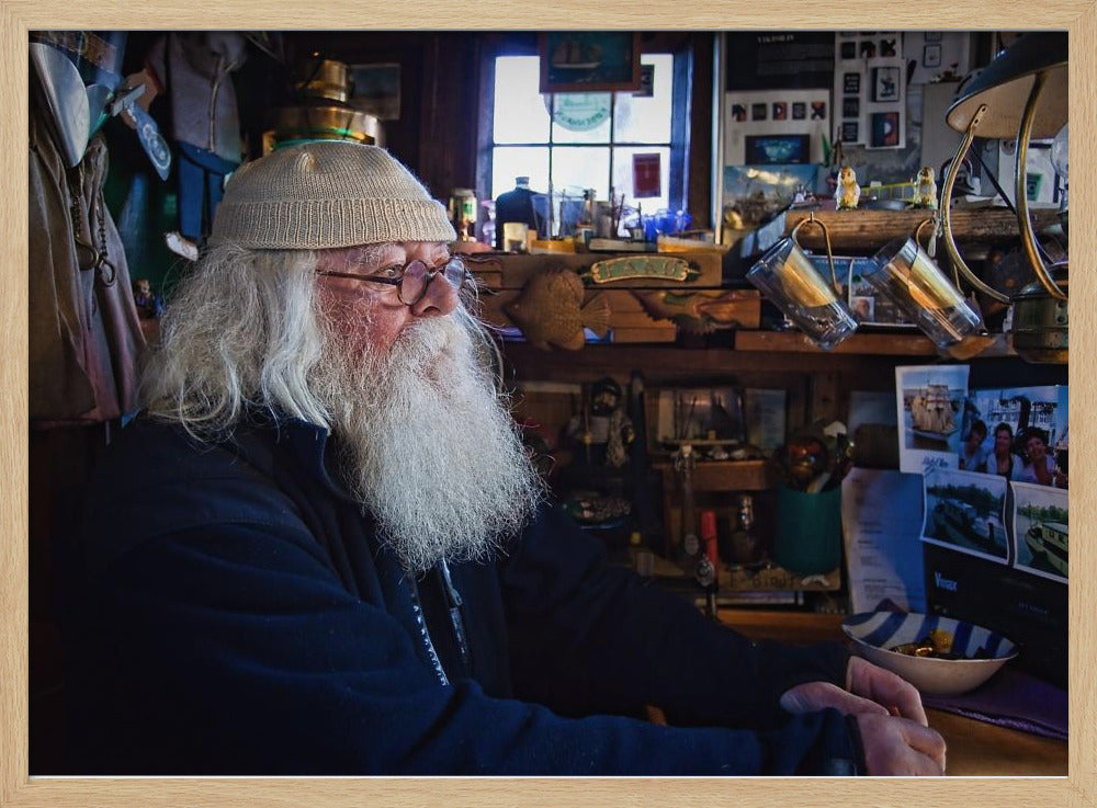 A profile portrait of an elderly man with a long, full white beard and hair, wearing glasses and a cream-colored knit beanie. He sits indoors in a warmly lit, cluttered room that looks like a cabin or a pub, with his hands clasped in front of him, gazing thoughtfully to the side. Poster