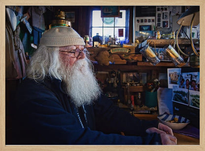 A profile portrait of an elderly man with a long, full white beard and hair, wearing glasses and a cream-colored knit beanie. He sits indoors in a warmly lit, cluttered room that looks like a cabin or a pub, with his hands clasped in front of him, gazing thoughtfully to the side. Poster
