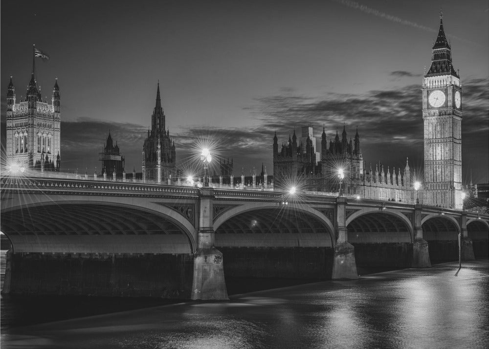 A framed, black and white nighttime photograph of Westminster Bridge in London. The iconic Houses of Parliament and the glowing Big Ben clock tower stand prominently in the background against a dark, cloudy sky. The streetlights on the bridge create starburst effects, and their reflections shimmer on the surface of the River Thames in the foreground. Poster