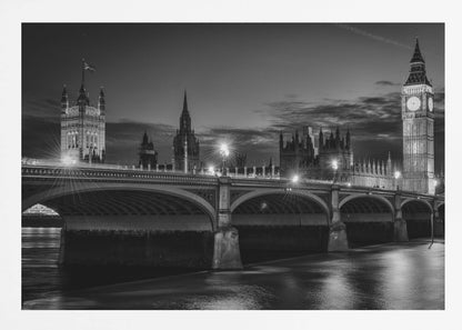A framed, black and white nighttime photograph of Westminster Bridge in London. The iconic Houses of Parliament and the glowing Big Ben clock tower stand prominently in the background against a dark, cloudy sky. The streetlights on the bridge create starburst effects, and their reflections shimmer on the surface of the River Thames in the foreground. Poster
