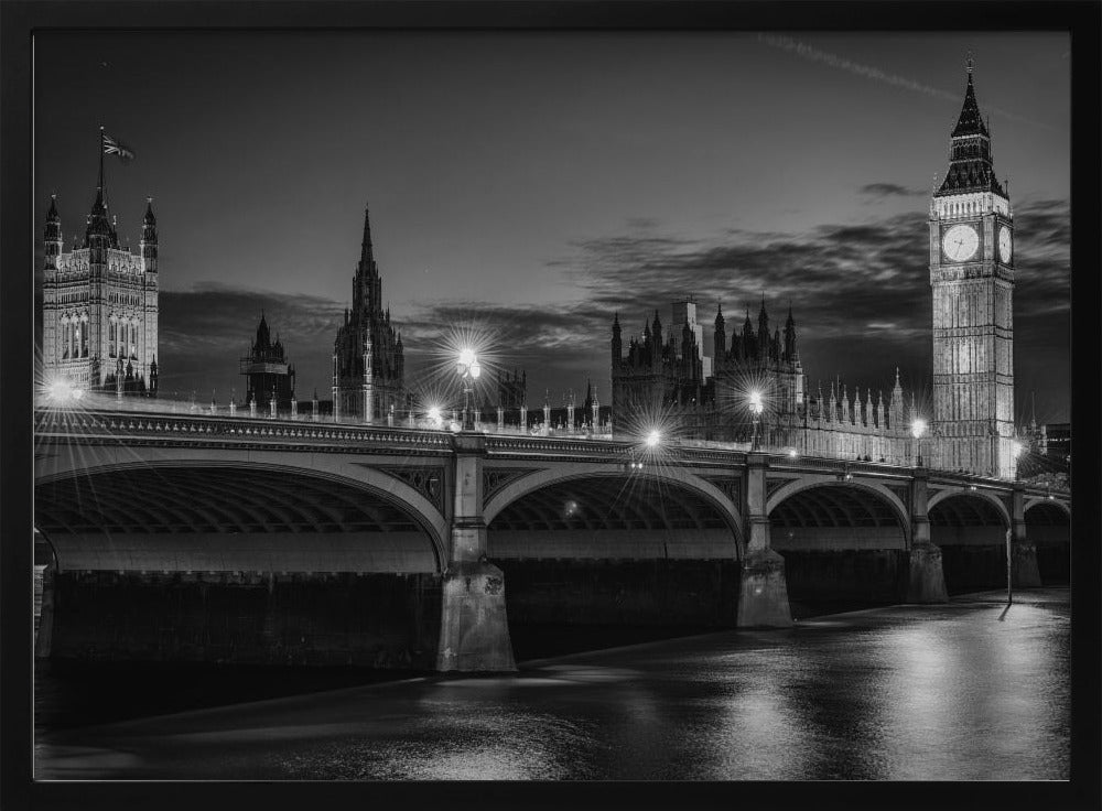 A framed, black and white nighttime photograph of Westminster Bridge in London. The iconic Houses of Parliament and the glowing Big Ben clock tower stand prominently in the background against a dark, cloudy sky. The streetlights on the bridge create starburst effects, and their reflections shimmer on the surface of the River Thames in the foreground. Poster