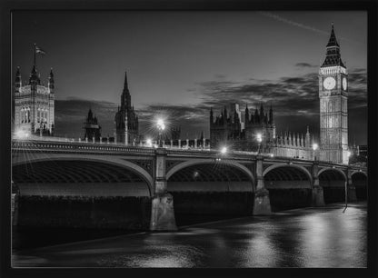 A framed, black and white nighttime photograph of Westminster Bridge in London. The iconic Houses of Parliament and the glowing Big Ben clock tower stand prominently in the background against a dark, cloudy sky. The streetlights on the bridge create starburst effects, and their reflections shimmer on the surface of the River Thames in the foreground. Poster